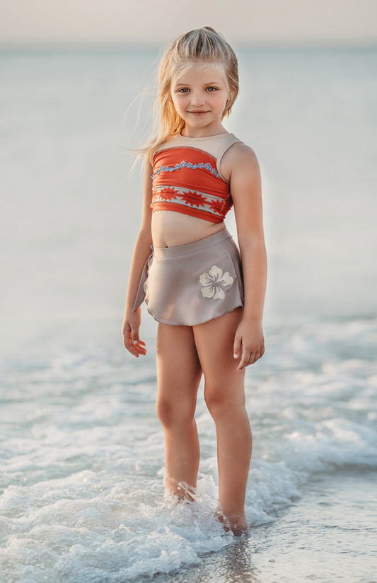 A young girl stands at the edge of the sea, smiling. She's wearing a bright red swim top with flower and pattern band.  The tan-grey swimsuit bottom is adorned with a tan hibiscus flower—it's the Polynesian Princess Two Piece Swimsuit made from SPF 50 material. The gentle waves touch her feet, blending with sky hues in the background.