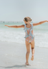 A young girl smiles brightly, wearing the Nora Leotard in Wildflower, adorned with a beautiful floral pattern and ruffles. The ruffles are light blue polka dot.  The wildflower body has pink, orange, blue green and light yellow. 