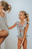 A young girl smiles brightly, wearing the Nora Leotard in Wildflower, adorned with a beautiful floral pattern and ruffles. The ruffles are light blue polka dot.  The wildflower body has pink, orange, blue green and light yellow. 