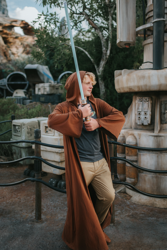 A young teen boy is wearing a sienna brown robe with hood wielding a light saber.  The robe is open and has large sleeves.  The robe ankle-floor length. 