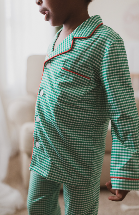 A young child is wearing the Gingham Plaid Unisex two-piece pajama set, featuring a green and white checkered pattern with red piping and white buttons. Ideal for Christmas family photos. 