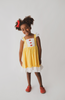 A young girl with long, wavy hair stands barefoot against a plain backdrop, wearing the Our Original Red Rose Twirl Dress from the Princess Twirl Collection. The sleeveless yellow dress features white ruffled trim and red buttons down the front. She holds the sides of her dress out slightly, creating a gentle arc.