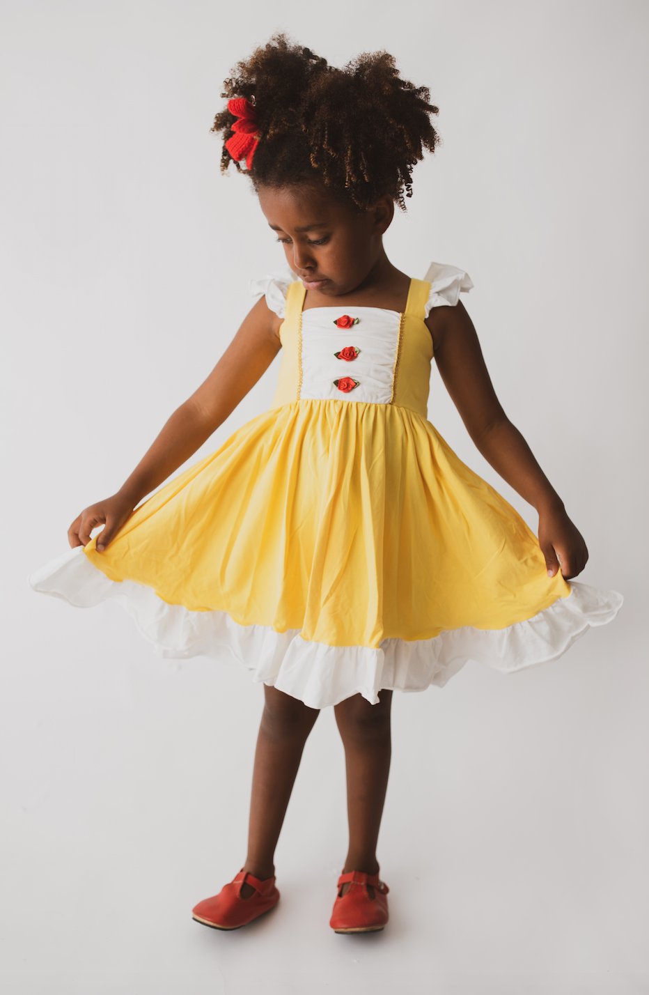 A young girl is wearing our Original Red Rose Twirl Dress from the Princess Twirl Collection. The sleeveless yellow dress features white ruffled trim and three red roses down the front. The sunny yellow dress provides a perfect princess twirl and soft comfortable cotton fabric. 