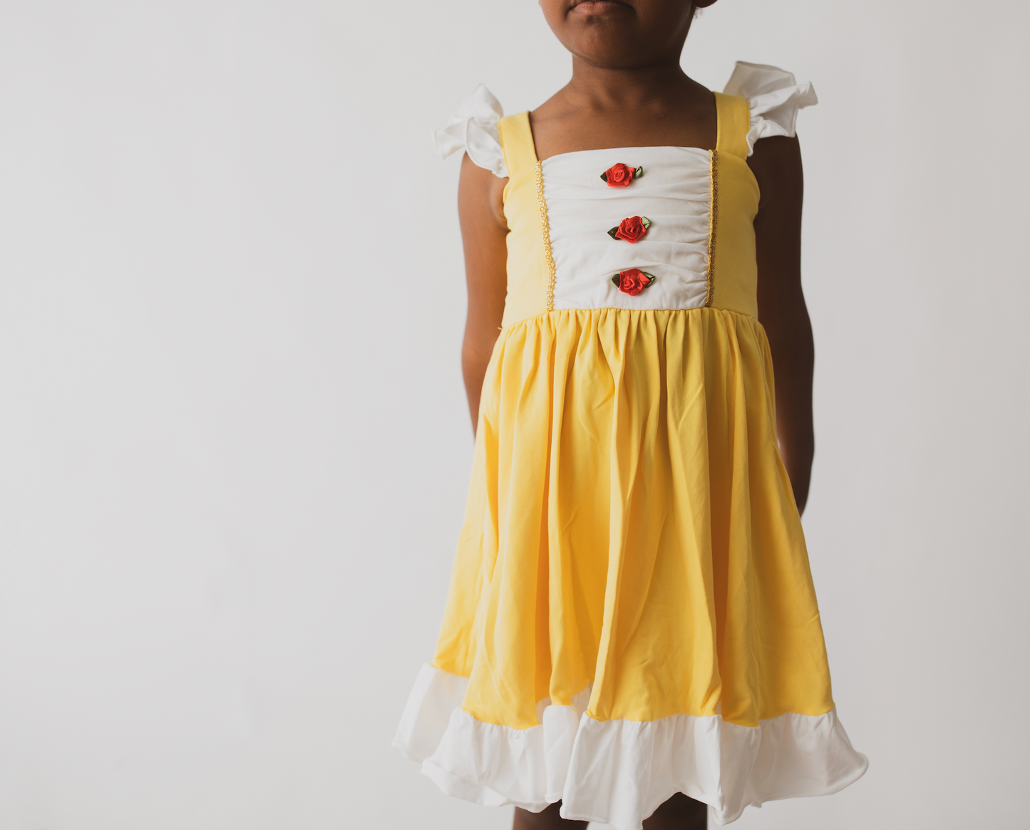 A young girl is wearing our Original Red Rose Twirl Dress from the Princess Twirl Collection. The sleeveless yellow dress features white ruffled trim and three red roses down the front. The sunny yellow dress provides a perfect princess twirl and soft comfortable cotton fabric. 