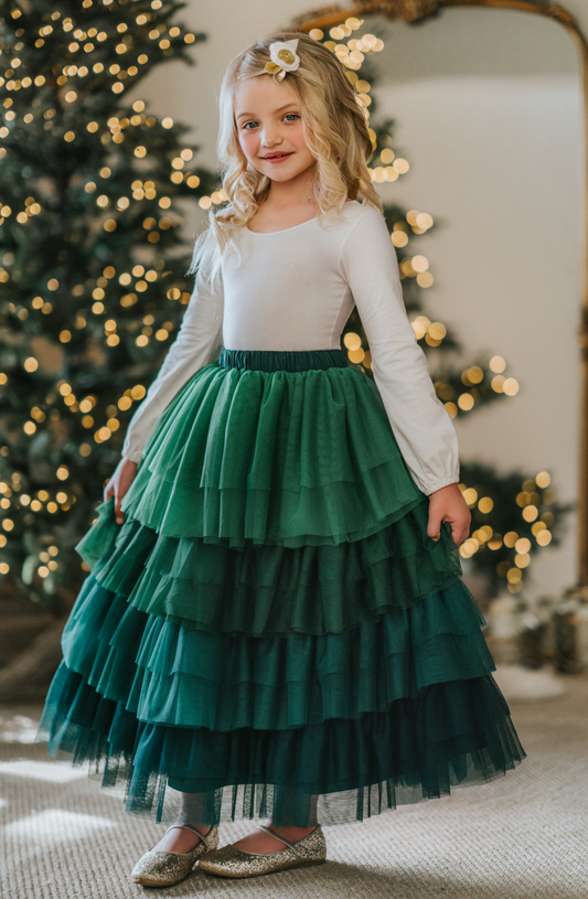 A young girl is wearing a long skirt in layers of green tulle.  The ruffles  cascade from green to evergreen.  There are 4 colors of green and 7 layers of tulle with a cotton liner for comfort. 