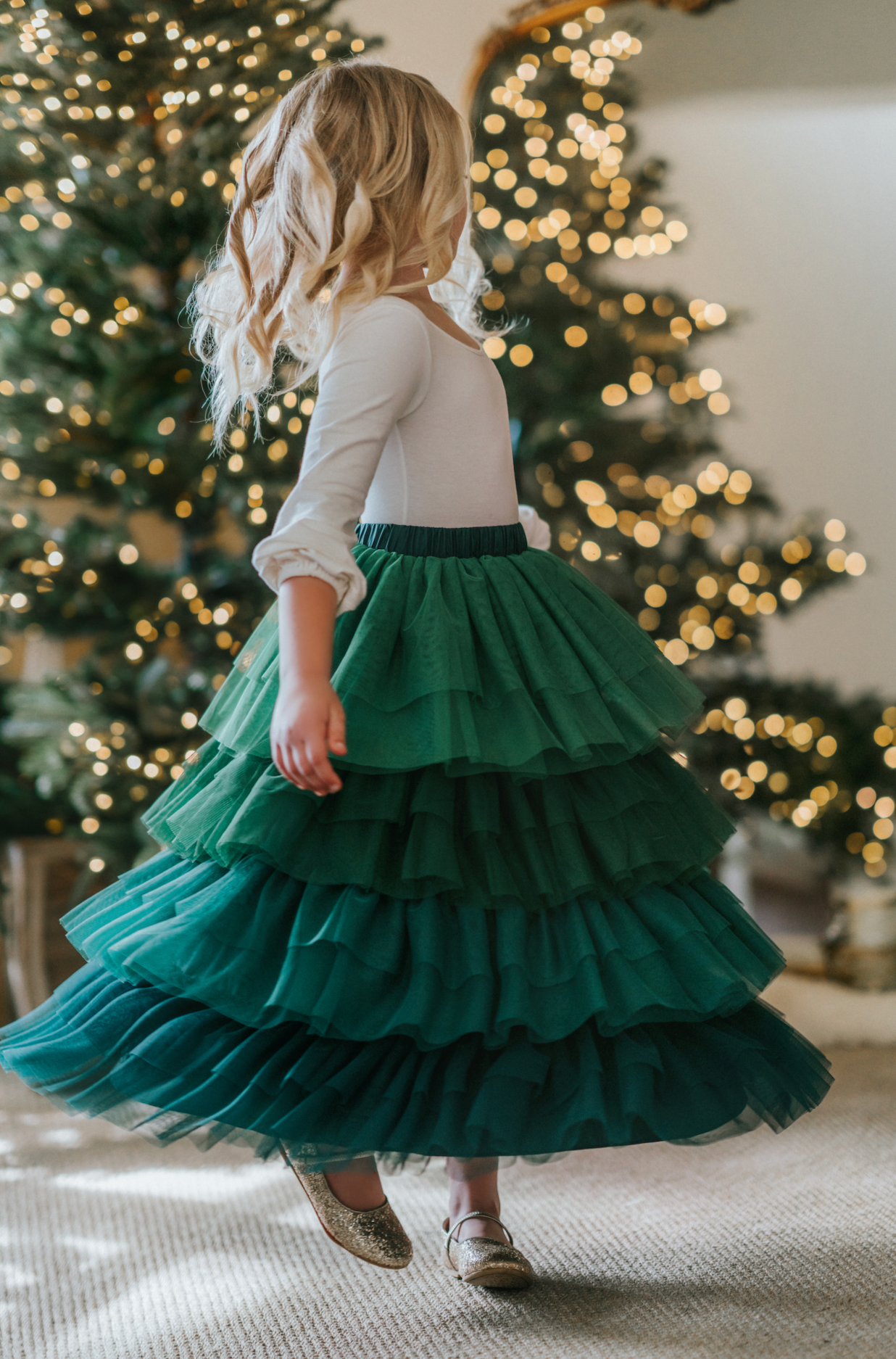 A young girl is wearing a long skirt in layers of green tulle.  The ruffles  cascade from green to evergreen.  There are 4 colors of green and 7 layers of tulle with a cotton liner for comfort. 