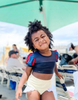 A young girl is wearing the Snow White  2 piece Swim Set with SPF 50 for sun protection.  The navy blue top has blue and red puff sleeves and navy mesh at the top.  The light yellow bottoms have a white ruffle on the leg. 