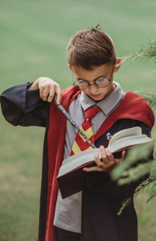 A young boy dressed in the Wizard Ensemble, featuring a red and yellow striped tie, and a black cotton jersey knit robe reminiscent of the Harry Potter series. The robe is lined with brick red and there is a logo sewn over the left chest. 