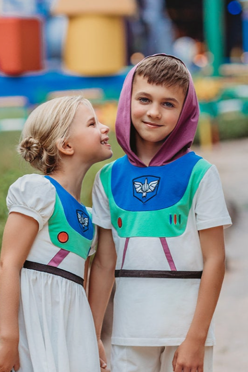 A young boy is wearing a purple hood attached to a white t-shirt.  There is blue and green on the chest of the shirt  with purple lines and a black band along the waist resembling an astronaut. There is an embroidered white symbol, red circle and red, blue and green lines that resemble space suit buttons. 