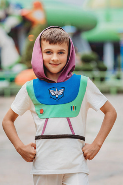 A young boy is wearing a purple hood attached to a white t-shirt.  There is blue and green on the chest of the shirt  with purple lines and a black band along the waist resembling an astronaut. There is an embroidered white symbol, red circle and red, blue and green lines that resemble space suit buttons. 