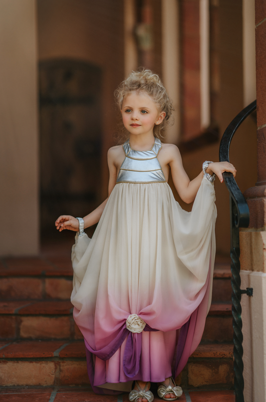 A young girl is dressed in a flowing white and purple gradient Portrait Collection Cosmic Queen Gown. The bodice and neck is silver with gold piping.  There are silver wristlets attached to the skirt of the gown and a ornate tie in the shape of a flower at the hem holding the skirts up. 