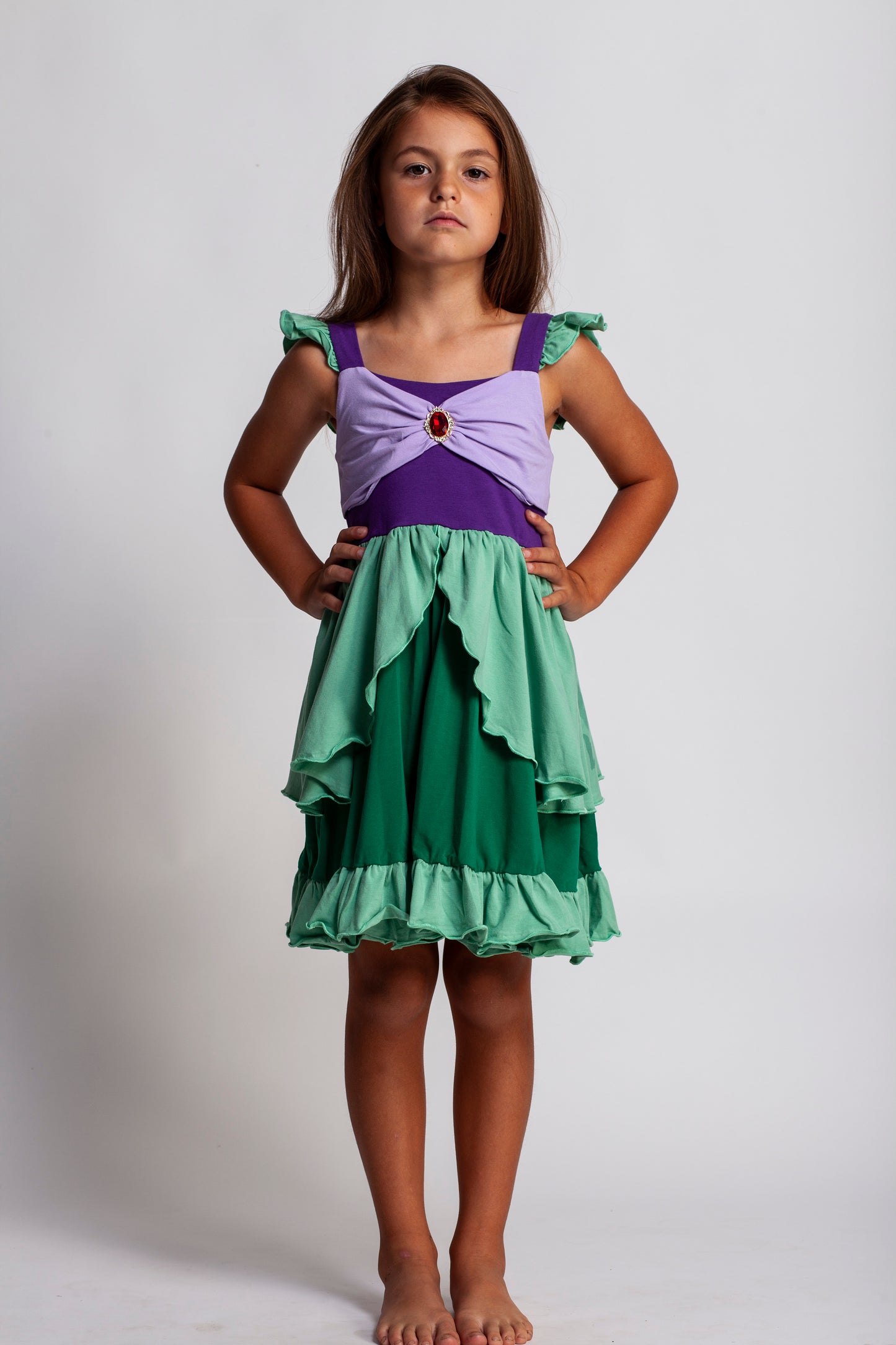 A young girl is wearing Our Original Mermaid Princess Twirl Dress in shades of green and purple. The sleeveless dress has green ruffle accent. The bodice has light and dark purple with a red ruby jewel. The twirl skirt is light and dark green with a ruffle at the hem. 