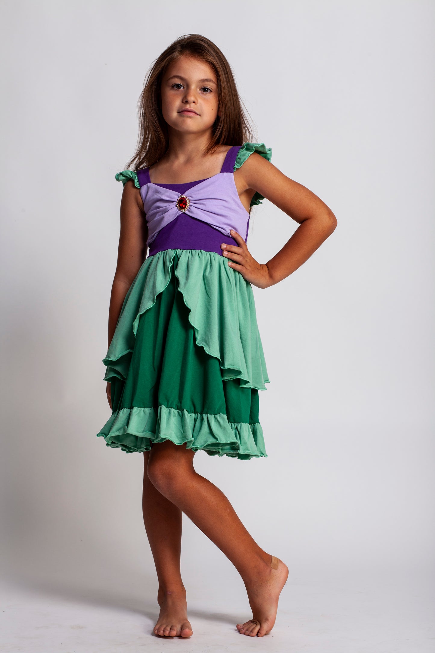 A young girl is wearing Our Original Mermaid Princess Twirl Dress in shades of green and purple. The sleeveless dress has green ruffle accent. The bodice has light and dark purple with a red ruby jewel. The twirl skirt is light and dark green with a ruffle at the hem. 