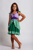 A young girl is wearing Our Original Mermaid Princess Twirl Dress in shades of green and purple. The sleeveless dress has green ruffle accent. The bodice has light and dark purple with a red ruby jewel. The twirl skirt is light and dark green with a ruffle at the hem. 