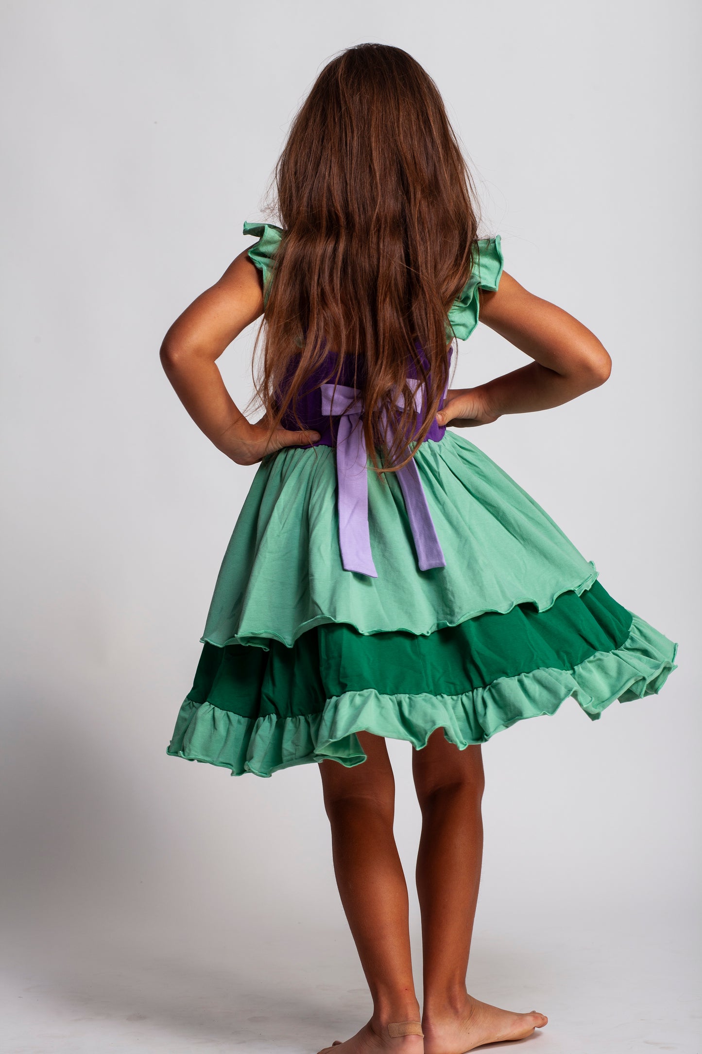 A young girl is wearing Our Original Mermaid Princess Twirl Dress in shades of green and purple. The sleeveless dress has green ruffle accent. The bodice has light and dark purple with a red ruby jewel. The twirl skirt is light and dark green with a ruffle at the hem. 