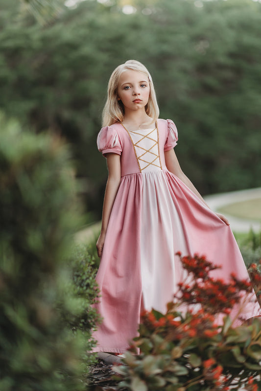 A young girl wearing a pink Sleeping Beauty Princess Maxi Dress. Short puff sleeves.  A lighter pink panel is on the chest with gold rick rack criss crossed accent over it. The long dress is a soft cotton. 