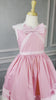 A young girl is wearing The Fairy Godmother Collection Cinderelly Pink Dress. The dress is woven cotton with a white collar and pink bow. The full skirt has bows at the bottom with a white hem. 