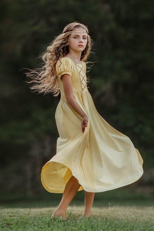 A young girl wearing a light yellow Princess Maxi Dress. Short puff sleeves.  A lighter yellow panel is on the chest with gold rick rack criss crossed accent over it. The long dress is a soft cotton. 