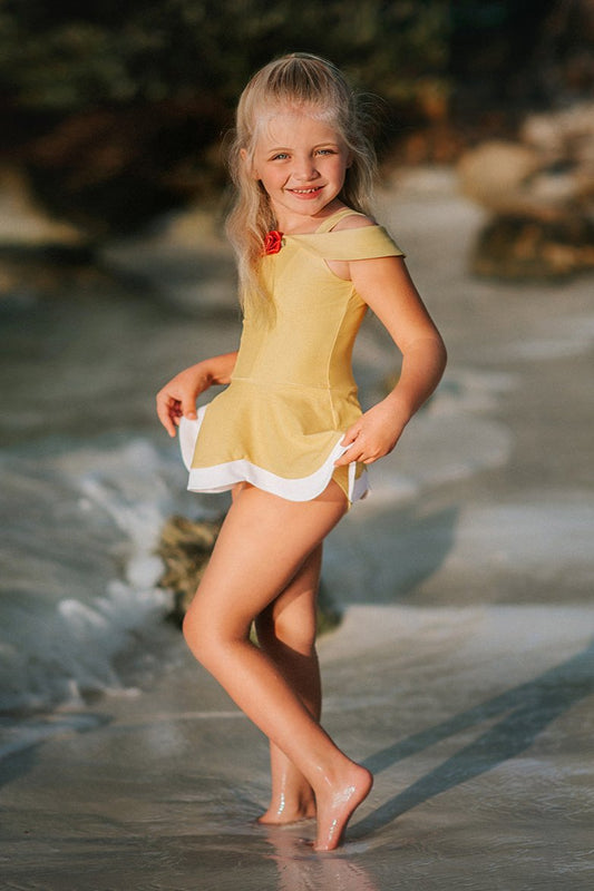 A young girl with long blonde hair is standing on the beach, wearing a yellow swimsuit with white trim and a red rose detail from the Red Rose Princess Collection. She is smiling and looking towards the camera with the ocean waves behind her. The setting appears to be sunny and warm.