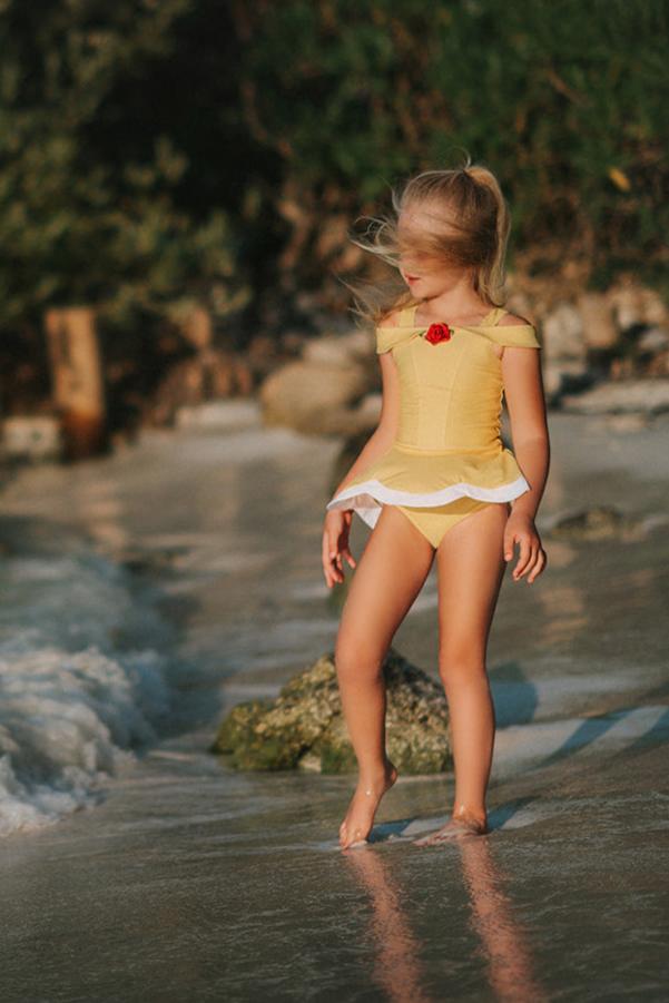 A young girl with long blonde hair is standing on the beach, wearing a yellow swimsuit with white trim and a red rose detail from the Red Rose Princess Collection. She is smiling and looking towards the camera with the ocean waves behind her. The setting appears to be sunny and warm.