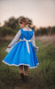 A young girl stands wearing the Red Rose Princess Village Dress from the Fairy Godmother Collection, reminiscent of Disney's Belle from "Beauty and the Beast." The white sleeves are long. The dress has a bight blue chest and skirt.  There is a white apron at the waist and white embroidery lace at the base. Dress is mid calf.
her 100% woven cotton dress to flare out. The background is softly blurred with trees and sky visible.