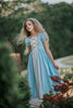 A young girl wearing a light blue Princess Maxi Dress. Short puff sleeves.  A lighter blue panel is on the chest with gold rick rack criss crossed accent over it. The long dress is a soft cotton. 