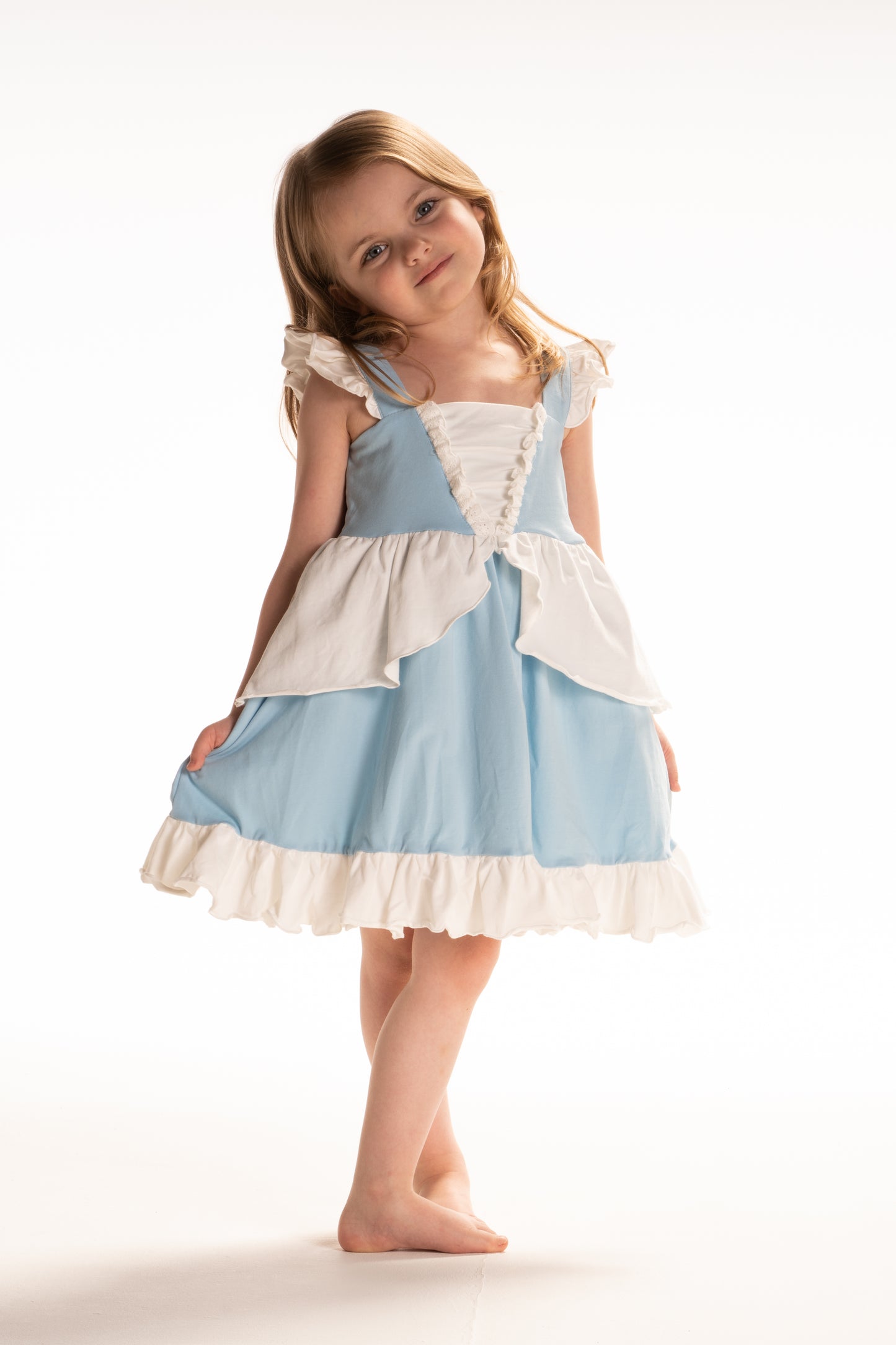 A young girl in an "Our Original Cinderella Twirl Dress" stands near a castle.  The dress has a white flutter ruffle on the straps. There is a white skirt ruffle that overlays the blue twirl skirt. The soft cotton material is comfortable and breathable. 