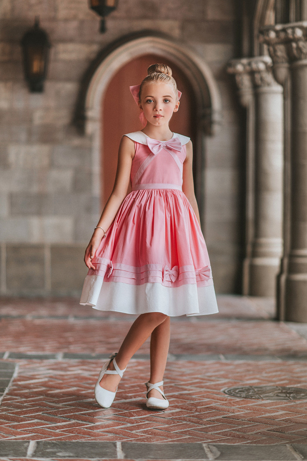 A young girl is wearing The Fairy Godmother Collection Cinderelly Pink Dress. The dress is woven cotton with a white collar and pink bow. The full skirt has bows at the bottom with a white hem. 