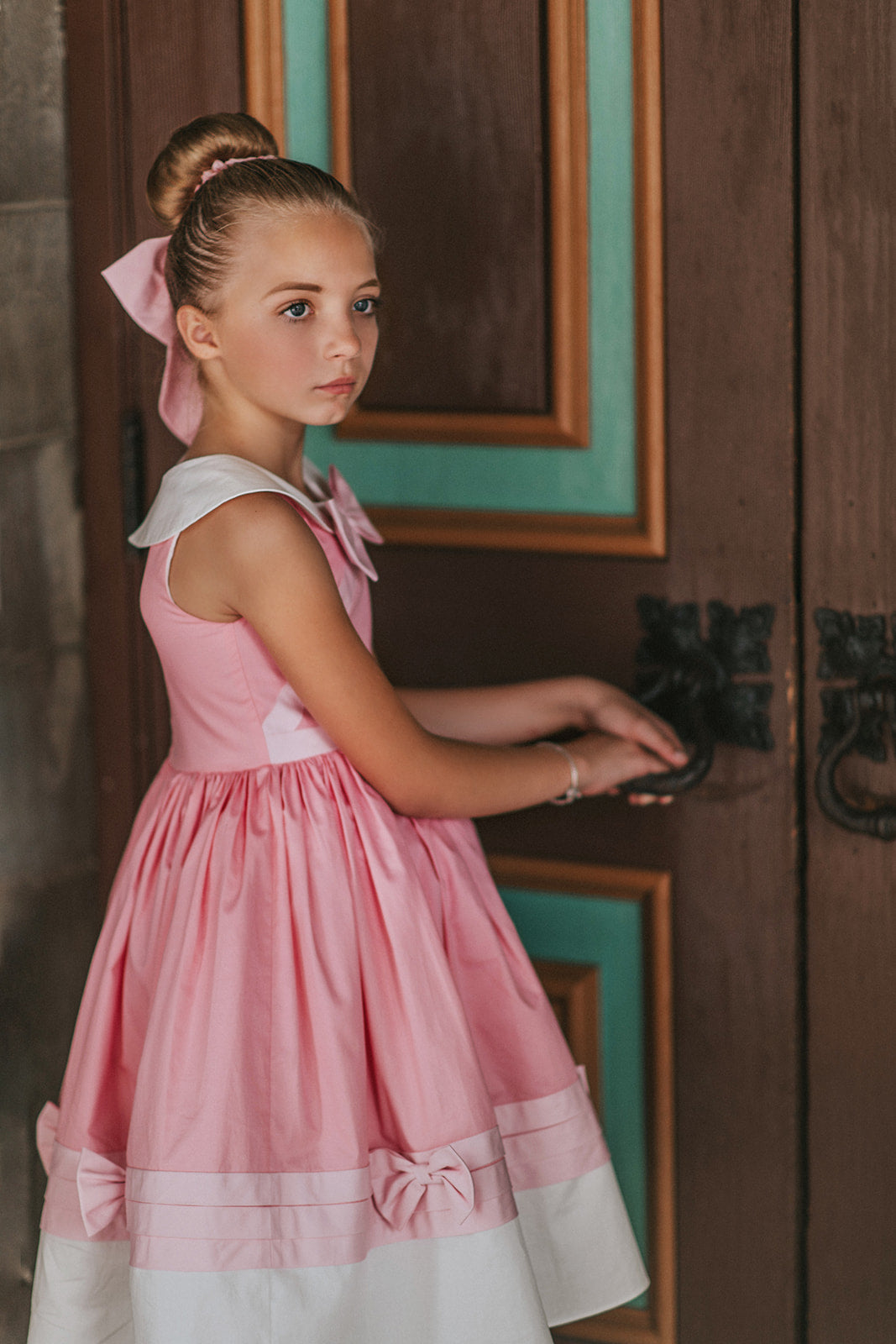 A young girl is wearing The Fairy Godmother Collection Cinderelly Pink Dress. The dress is woven cotton with a white collar and pink bow. The full skirt has bows at the bottom with a white hem. 