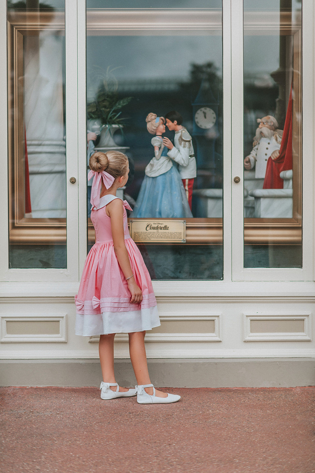 A young girl is wearing The Fairy Godmother Collection Cinderelly Pink Dress. The dress is woven cotton with a white collar and pink bow. The full skirt has bows at the bottom with a white hem. 