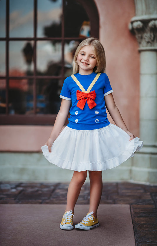 A young girl is wearing a blue tshirt with sailor adornments on it.  The yellow v design has a red bow at the middle.  The white buttons and white trim sleeves make the perfect duck sailor outfit. 