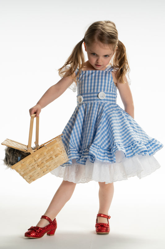 A young girl is wearing a blue  and white checkered dress. The sleeveless dress has white lace flutter details on the straps.  There are two white buttons sewn at the waist. At the bottom of the dress there is a white sewn in pettiskirt with eyelet detail.