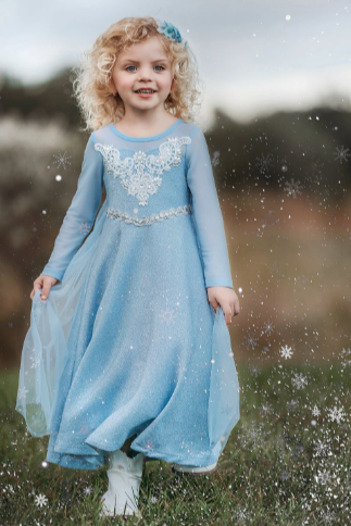 A young girl is dressed in the sparkly light blue Portrait Collection Ice Queen Gown adorned with intricate custom  lace at the neckline with rhinestones. A light blue cape with silver sparkle snowflakes is buttoned to the back of the gown. 