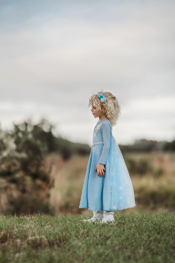 A young girl is dressed in the sparkly light blue Portrait Collection Ice Queen Gown adorned with intricate custom  lace at the neckline with rhinestones. A light blue cape with silver sparkle snowflakes is buttoned to the back of the gown. 