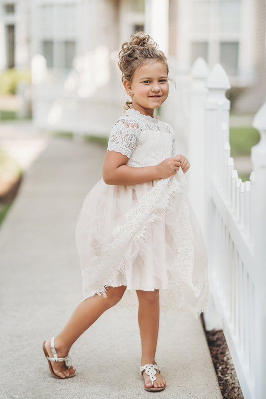 A young girl with curly hair is wearing an elegant Victorian Day Gown in Ivory and white sandals, standing on a sidewalk next to a white picket fence. She is smiling and holding the tulle skirt of her gown, with a residential area in the blurred background.