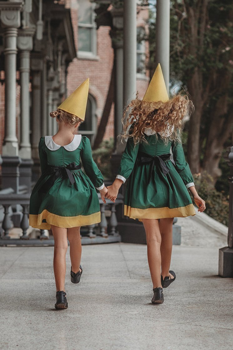 A young girl stands wearing the Smiling Elf Dress, adorned with yellow embroidery, a white collar, and a yellow hem. She also sports a matching yellow cone hat.