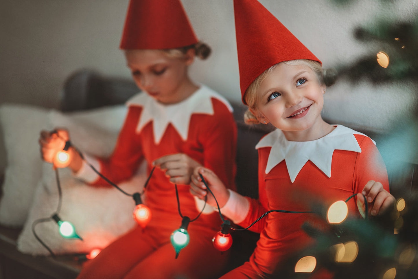 A young child dressed in Unisex Elf Pajamas with a red pointy hat. The perfect Christmas photo outfit. The fabric is bright red and buttery soft.  The top is long sleeved with white cuffs and a white collar that has large triangle points similar to a clown. Long pants with white cuffs. 