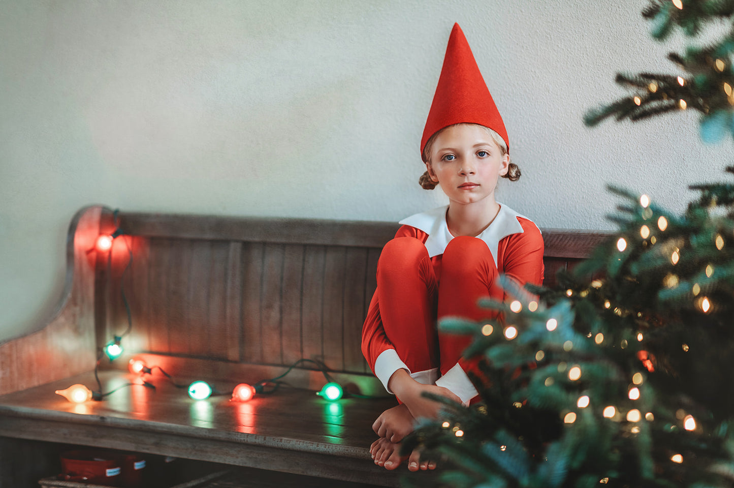 A young child dressed in Unisex Elf Pajamas with a red pointy hat. The perfect Christmas photo outfit. The fabric is bright red and buttery soft.  The top is long sleeved with white cuffs and a white collar that has large triangle points similar to a clown. Long pants with white cuffs. 