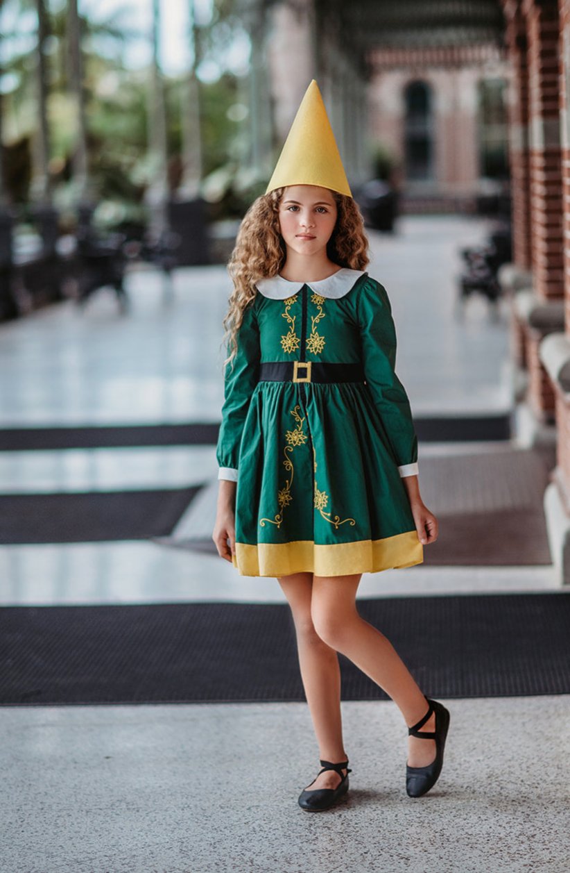 A young girl stands wearing the Smiling Elf Dress, adorned with yellow embroidery, a white collar, and a yellow hem. She also sports a matching yellow cone hat.