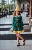 A young girl stands wearing the Smiling Elf Dress, adorned with yellow embroidery, a white collar, and a yellow hem. She also sports a matching yellow cone hat.