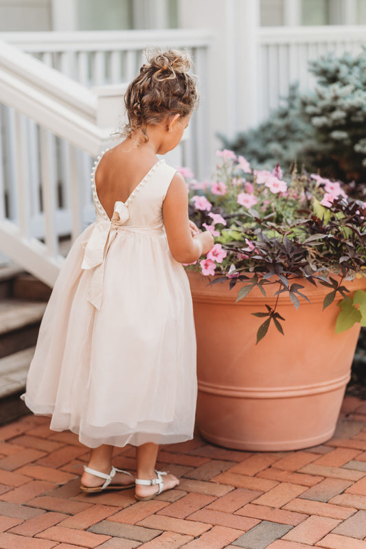 A young girl in a Pearl Accent gown in Antique Ivory, featuring a bow at the back. Woven cotton bodice and large pearl beads line the neck and back edge! A full tulle skirt with tea length hemline and modest open back.