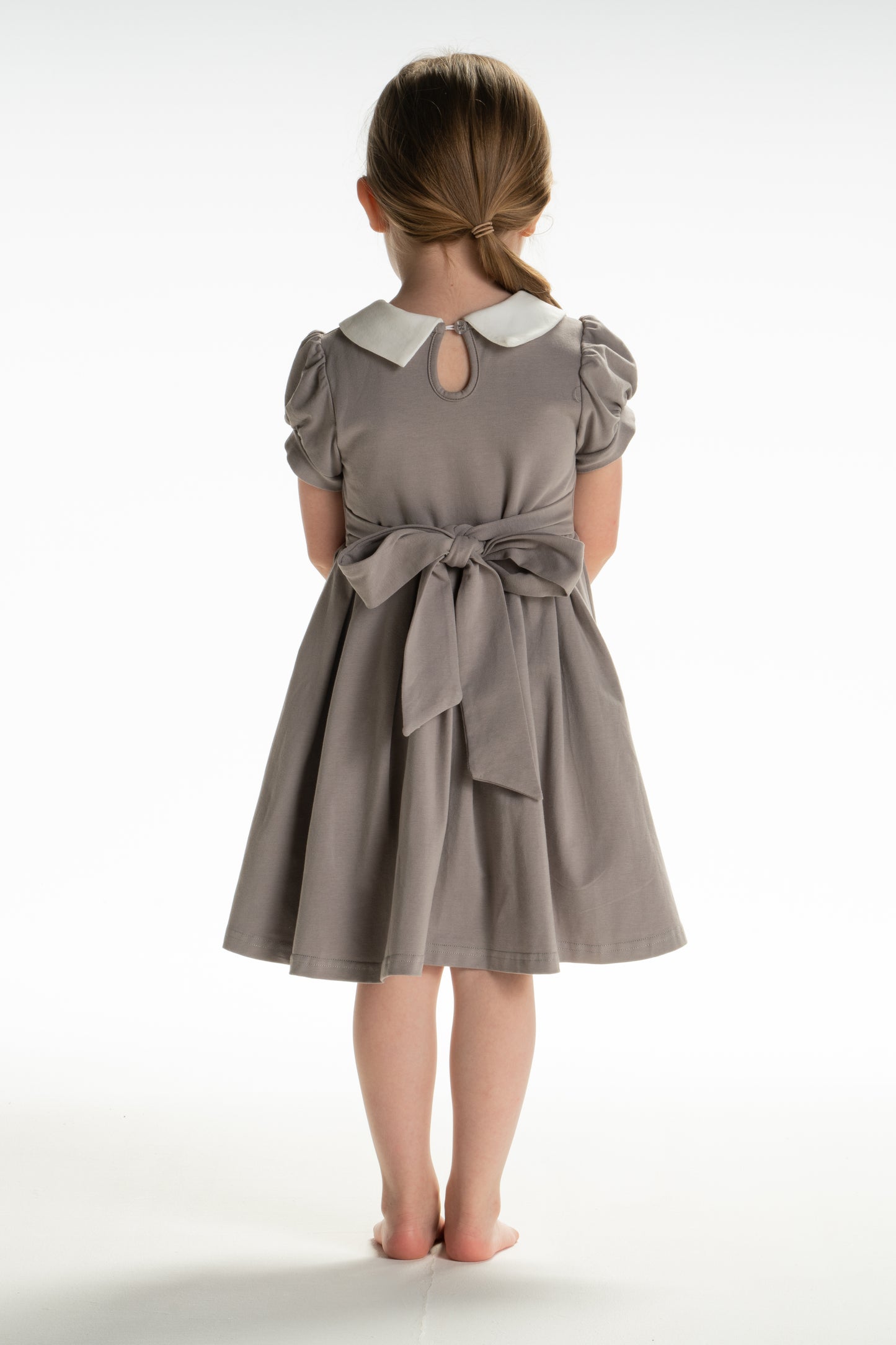 A young girl is wearing a grey Wizard Girl Twirl Dress with a striped yellow and brick red tie. The twirl dress has a white collar, short puff sleeves and a tie in the back at the waist. 