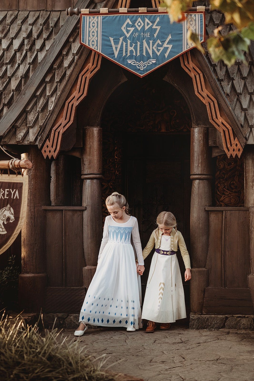 A young girl with blonde hair stands on a rock, wearing the Queen of the Forest Maxi Dress adorned with blue diamond patterns along the bottom. The maxi dress also features a geometric design on the upper part. She is looking downward and appears thoughtful, with a background of trees and buildings.