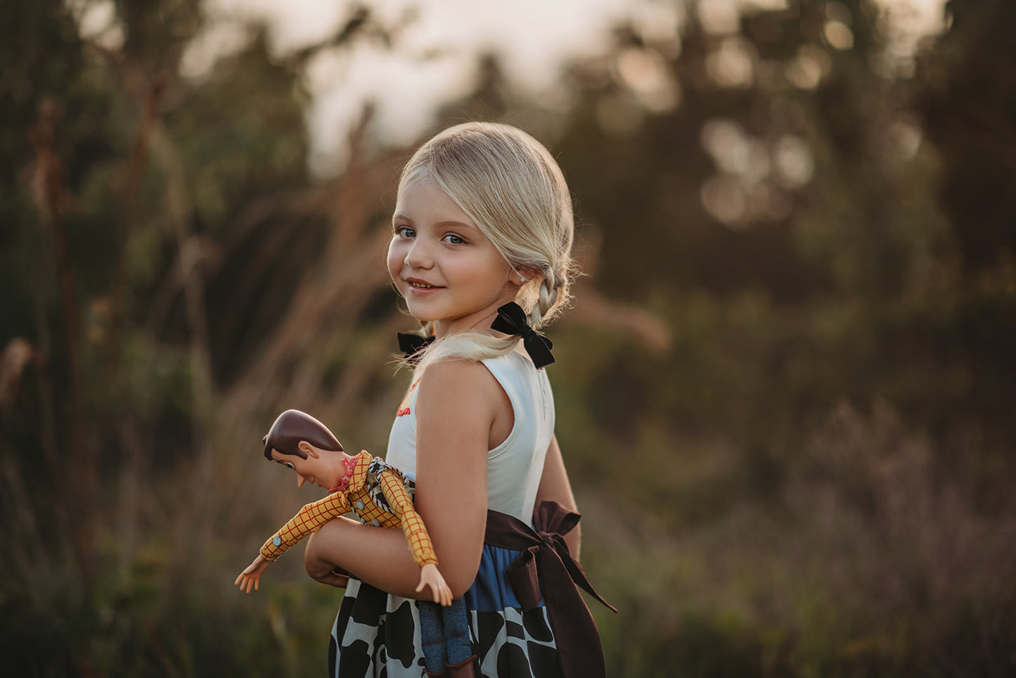 A young girl is wearing a Cowgirl Character Twirl Dress from the Toyland Collection. The sleeveless dress has a yellow and white bodice with red rick rack swirled across it.  The twirl skirt features a blue faux denim with black and white cow print and red rick rack trim. 