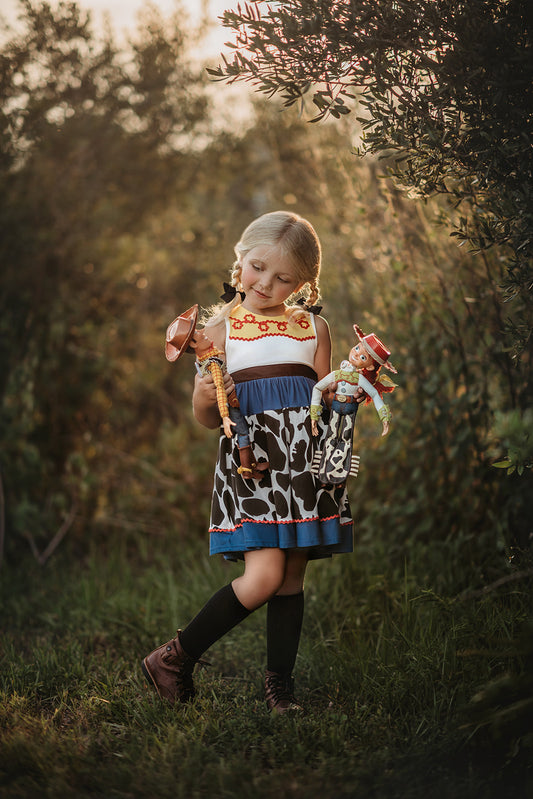 A young girl is wearing a Cowgirl Character Twirl Dress from the Toyland Collection. The sleeveless dress has a yellow and white bodice with red rick rack swirled across it.  The twirl skirt features a blue faux denim with black and white cow print and red rick rack trim. 