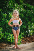 A young girl is wearing a 2 piece swim set.  The top is white with yellow on the upper chest.  Across the yellow is red scrolling rick rack.  The bottoms are a faux denim with black and white cow print panels. 