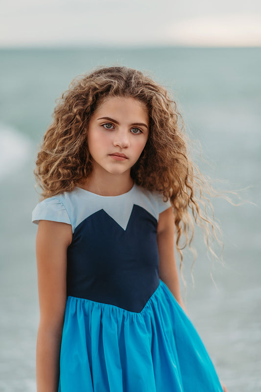 A young girl is wearing a Kiss the Girl Mermaid Dress from The Fairy Godmother Collection, featuring a navy bodice and light blue cap sleeves. The dress skirt is a bight cerulean blue. The material is woven cotton. 