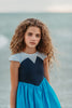 A young girl is wearing a Kiss the Girl Mermaid Dress from The Fairy Godmother Collection, featuring a navy bodice and light blue cap sleeves. The dress skirt is a bight cerulean blue. The material is woven cotton. 