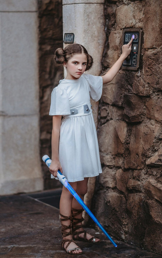 A young girl is wearing a white cotton twirl dress.  The dress has a white hood and short belle sleeves.  There are metallic silver designs and buttons on the waist and a tie in the back. 