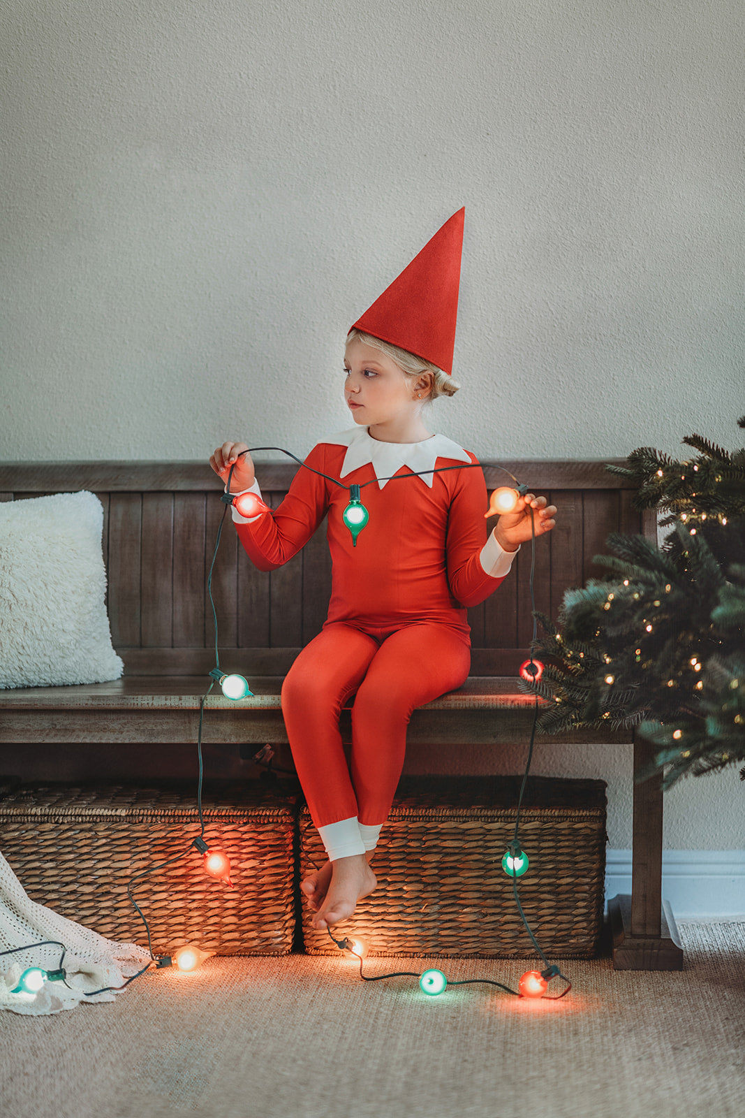 A young child dressed in Unisex Elf Pajamas with a red pointy hat. The perfect Christmas photo outfit. The fabric is bright red and buttery soft.  The top is long sleeved with white cuffs and a white collar that has large triangle points similar to a clown. Long pants with white cuffs. 